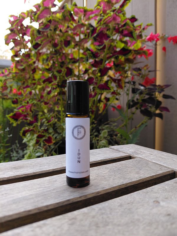 Sweet Aromatherapy Essential Oil by The Flower Pot in a sleek amber bottle on a wooden surface, with colorful garden foliage in the background. An ideal natural fragrance for relaxation and wellness.