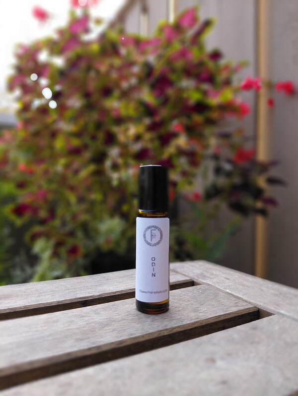 Essential oil bottle labeled "Odina" placed on a wooden surface with a blurred colorful flower pot in the background.