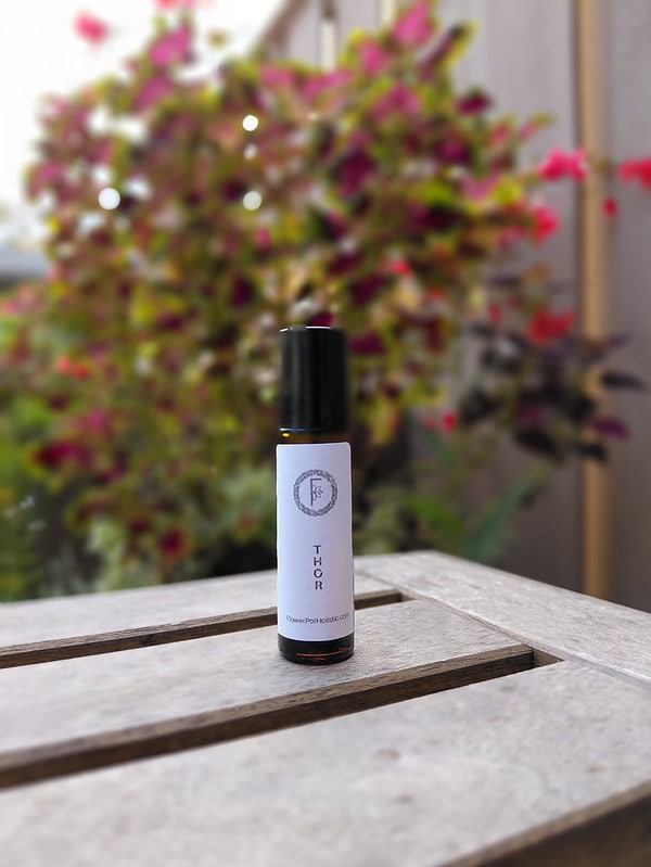 Aromatherapy essential oil bottle with "ThOR" label on wooden surface with colorful flowering plant background.